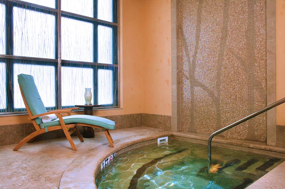 Spa relaxation area with wooden lounge chair beside a heated mineral hot tub with mosaic tiles