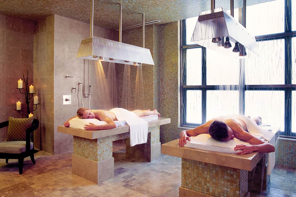 Luxurious spa treatment room with two massage tables, overhead rain showers, and ambient candlelight