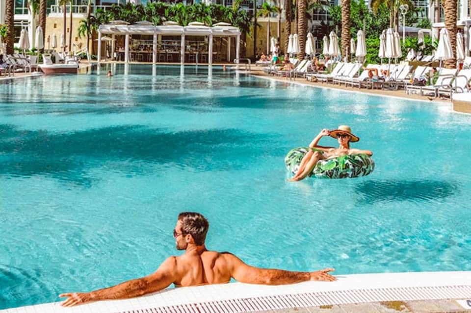 Man relaxing at edge of large resort pool with inflatable floats and lounge chairs