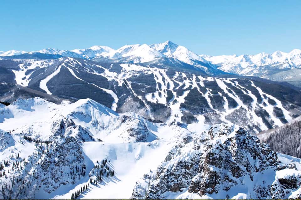 Snowy ski resort landscape with multiple ski runs, forested slopes, and snow-capped mountains in distance