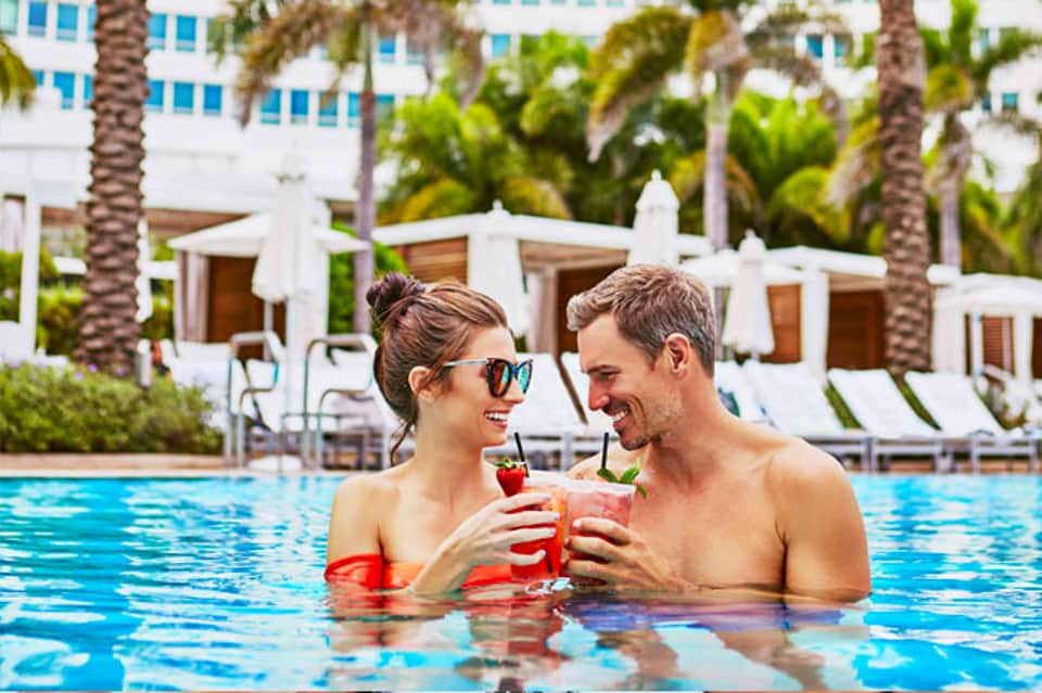 Couple enjoying tropical cocktails in resort pool with palm trees and white cabanas in background