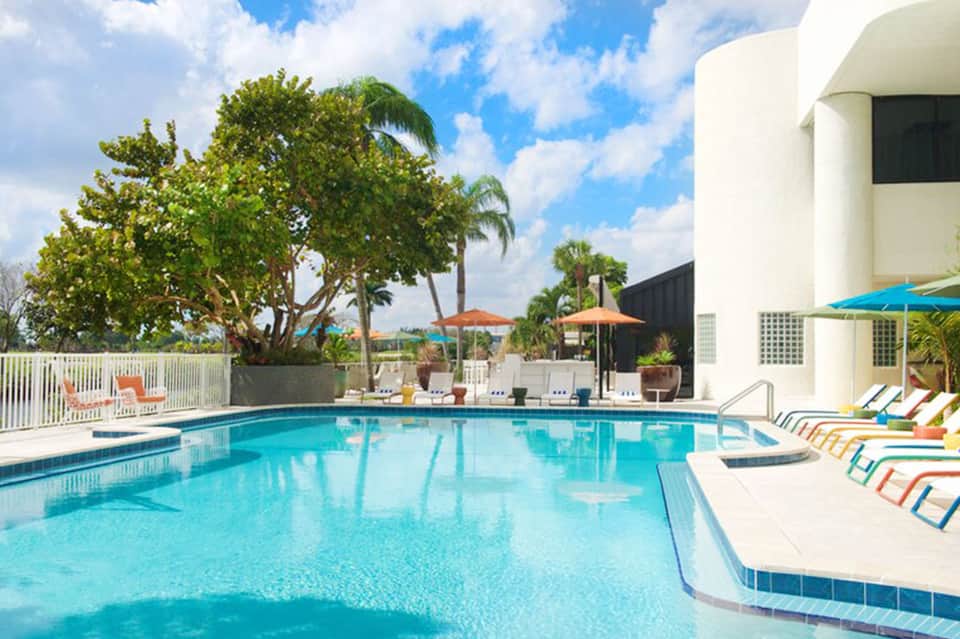 Curved swimming pool with colorful lounge chairs, white fence, palm trees, and blue umbrellas