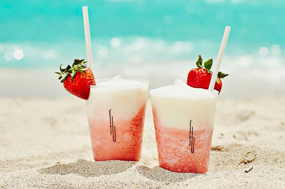 Two strawberry daiquiri cocktails with white straws on sand beach with turquoise ocean background