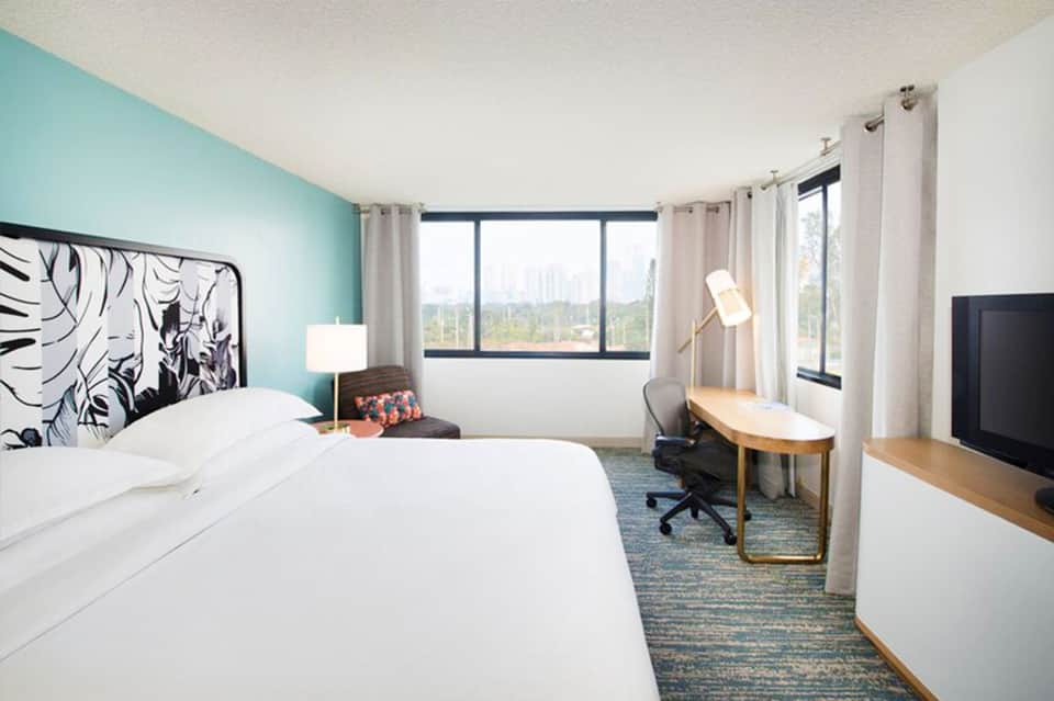 Modern hotel bedroom with turquoise accent wall, two queen beds, desk, and floor-to-ceiling windows with city views