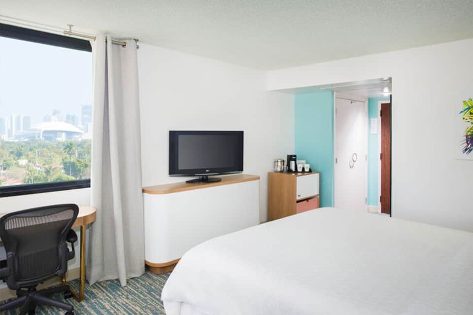 Modern hotel room with white bedding, TV, desk, and city skyline view through window