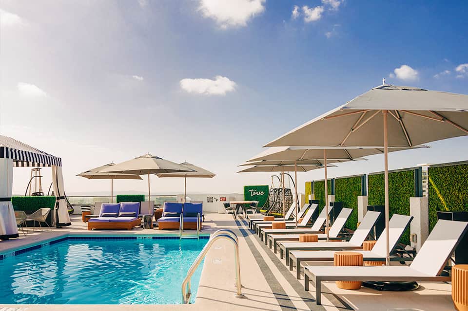 Resort rooftop pool with multiple umbrellas, loungers, and water features under clear blue sky