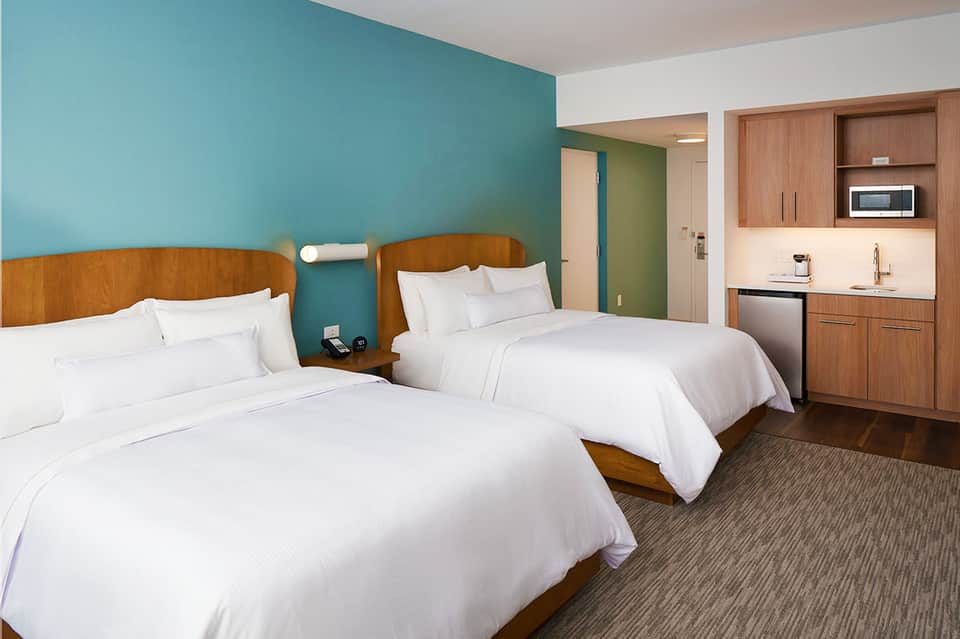 Modern hotel room with two queen beds, turquoise accent wall, wooden headboards, and kitchenette with wood cabinetry