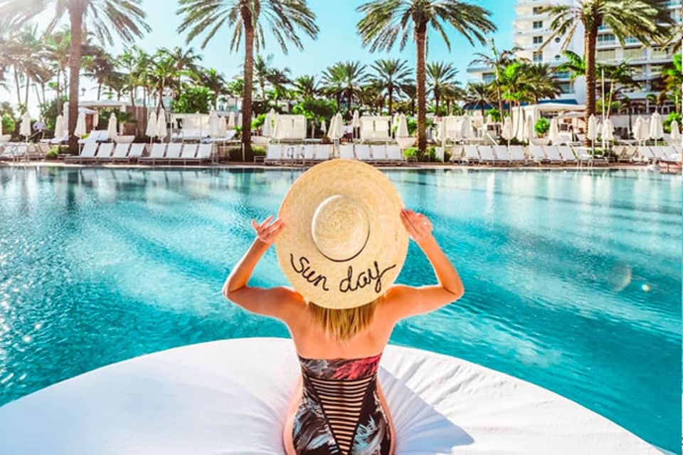 Woman in sun hat relaxing on lounge chair overlooking palm-lined resort pool