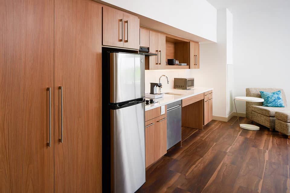 Modern hotel room kitchenette with wood cabinets, stainless steel appliances, and seating