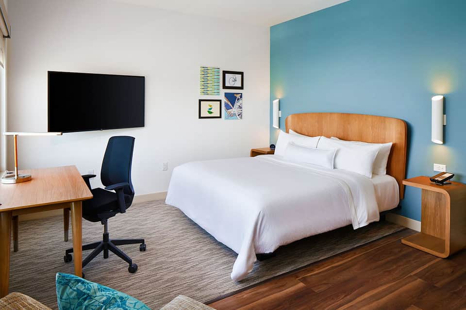 Modern hotel bedroom with blue accent wall, wooden bed frame, work desk, and flat-screen TV