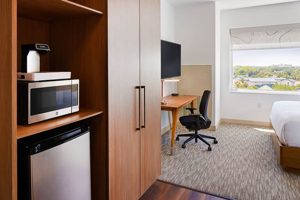 Modern hotel room with wood furnishings, kitchenette, work desk, and window overlooking suburban landscape