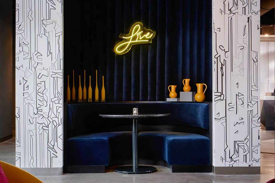 Modern hotel lounge with blue velvet seating, neon yellow 'live' sign, and decorative gold vessels on black shelving