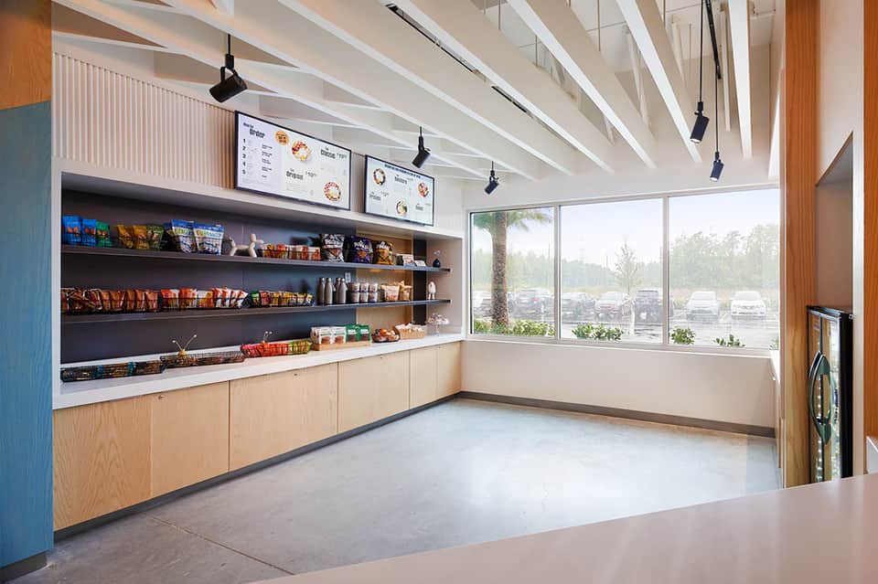 Contemporary café interior with wooden cabinetry, shelving with products, white ceiling beams, and large windows.