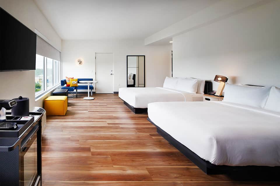 Modern hotel room with two queen beds, wooden floors, blue sofa, and floor-to-ceiling windows