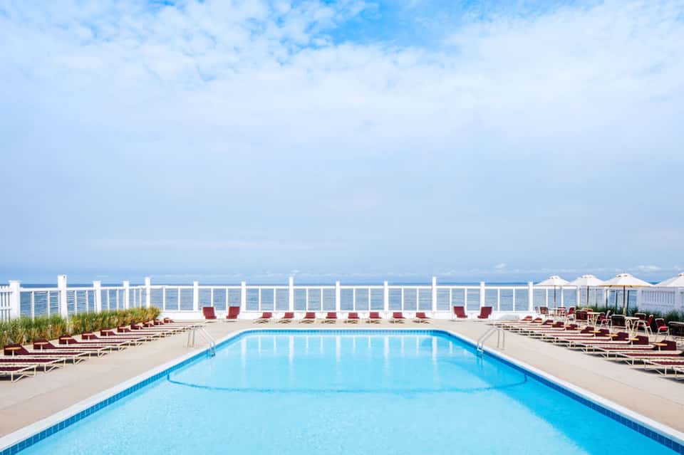 Oceanfront rectangular pool with white railings, burgundy loungers, and coastal views on clear day