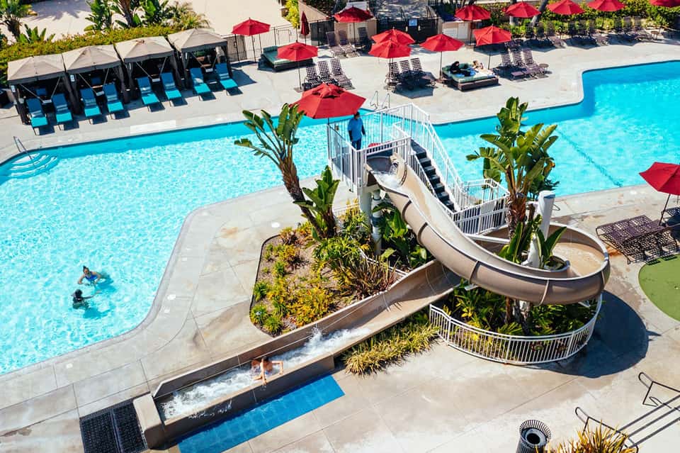 Resort pool with water slide, red umbrellas, lounge chairs, and tropical landscaping