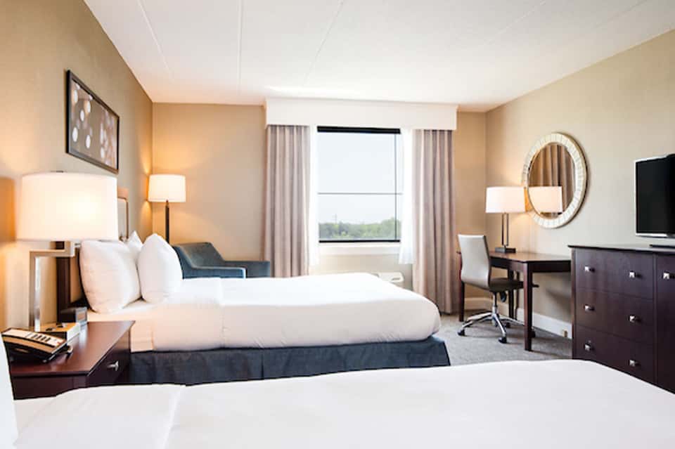 Modern hotel bedroom with two queen beds, work desk, and large windows overlooking green landscape