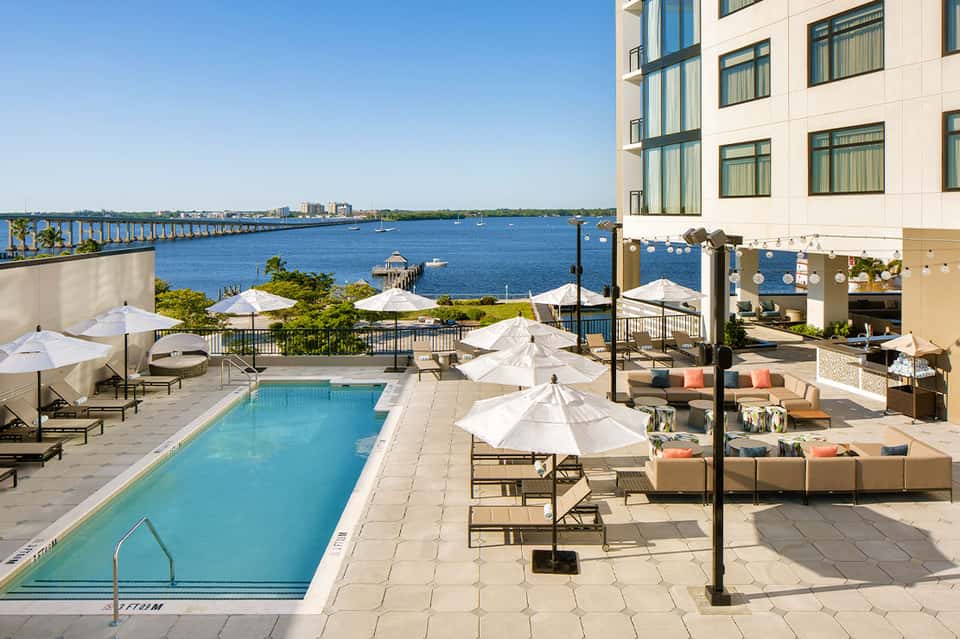 Waterfront hotel pool deck with white umbrellas, lounge chairs, and blue bay view