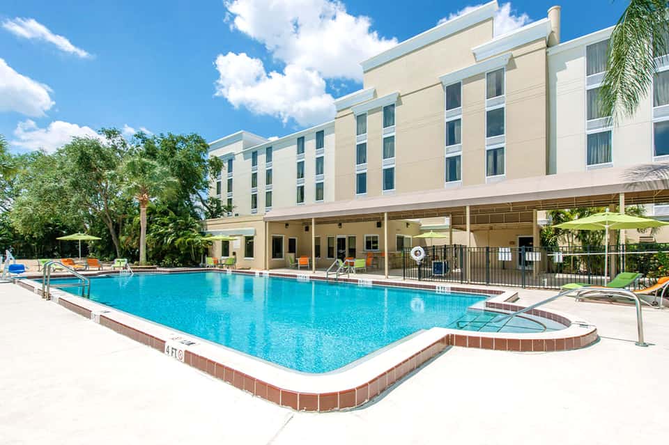 Large outdoor swimming pool with spa tub at hotel courtyard, surrounded by multi-story beige building and palm trees