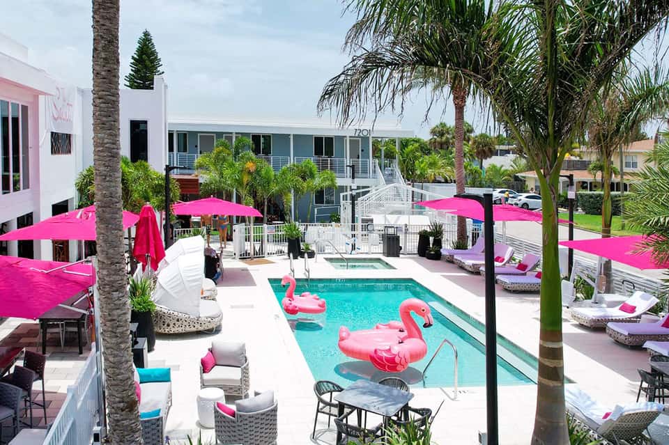 Bright pink umbrellas and flamingo floats surround a turquoise pool at a modern white resort with palm trees