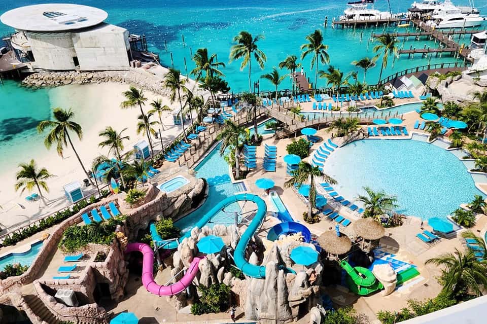 Aerial view of large tropical resort with multiple pools, water slides, blue umbrellas, and turquoise ocean