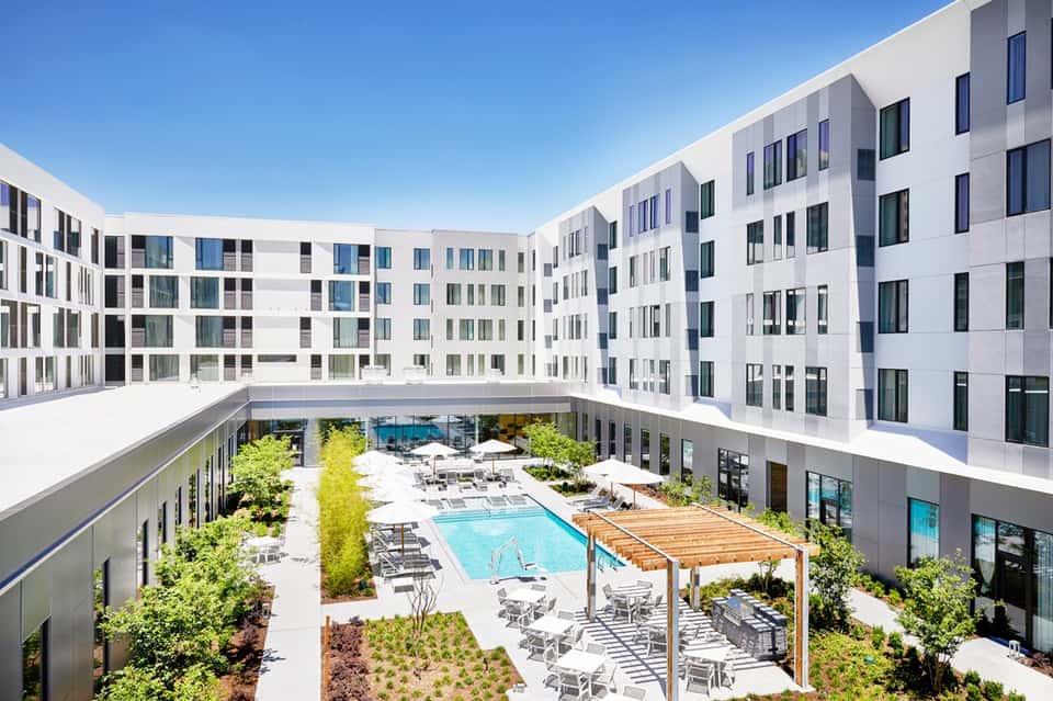 Modern white apartment complex courtyard with swimming pool, lounge chairs, wooden pergola, and surrounding greenery
