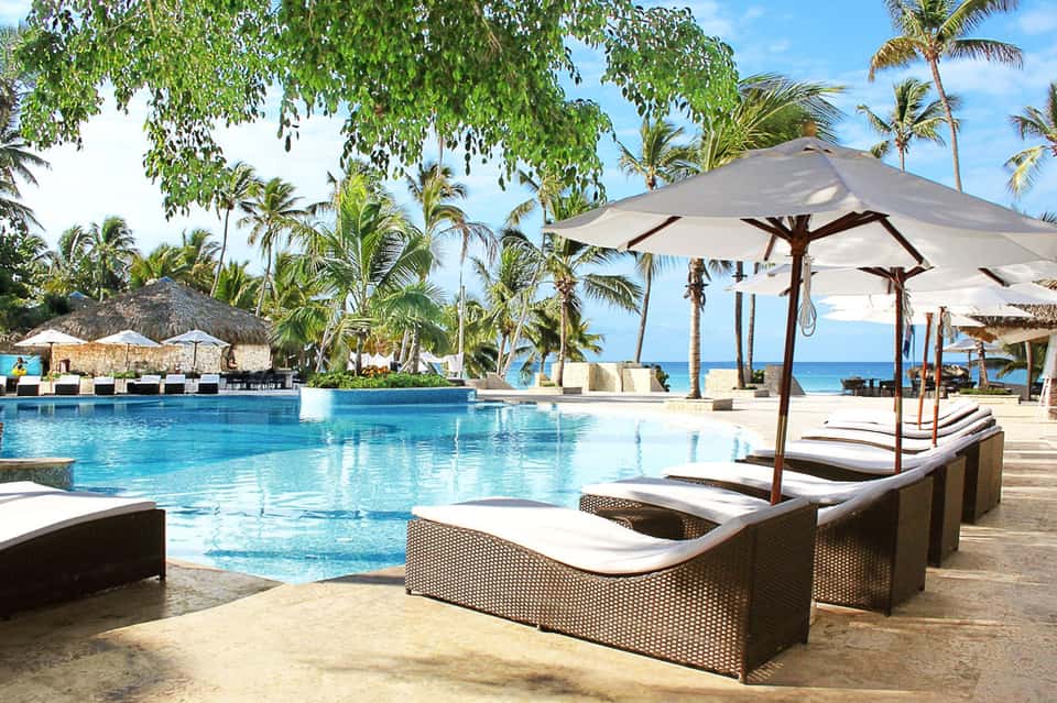 Beachfront resort pool with turquoise water, palm trees, white umbrellas, and ocean in background