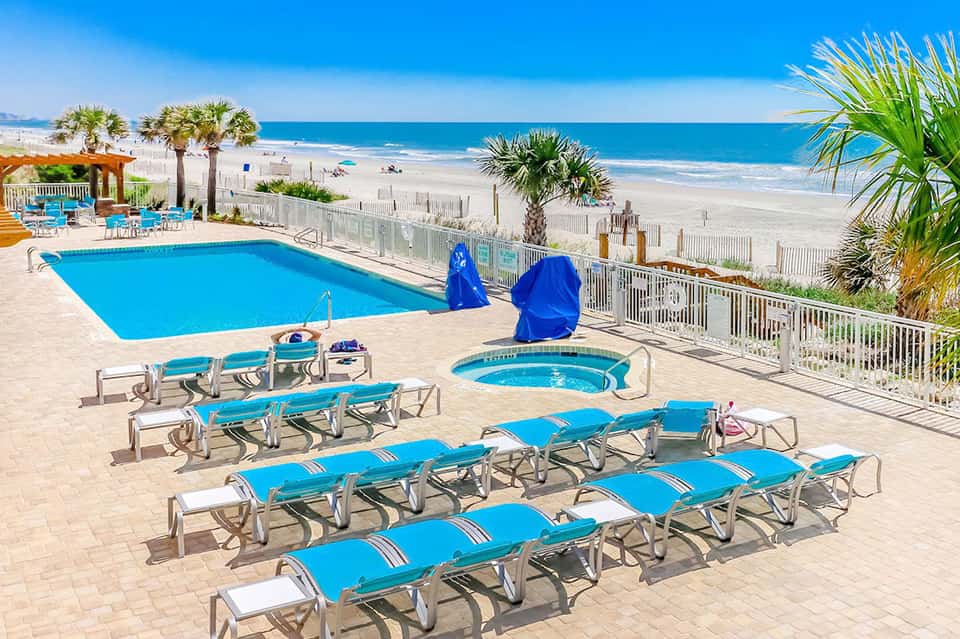 Beachfront pool with blue loungers, umbrellas, hot tub, and ocean view beyond white fence