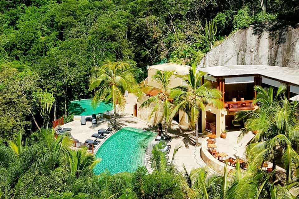 Aerial view of luxury resort with turquoise pools, palm trees, and wooden structures nestled in lush jungle vegetation