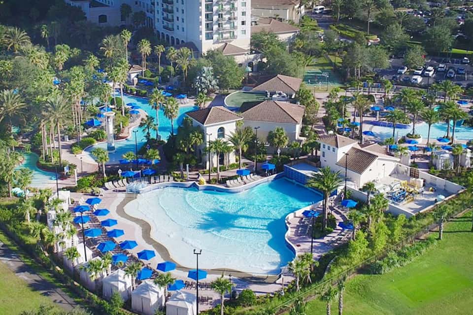 Aerial view of resort with curved blue pool, palm trees, white buildings, and lounging areas surrounded by manicured lawns