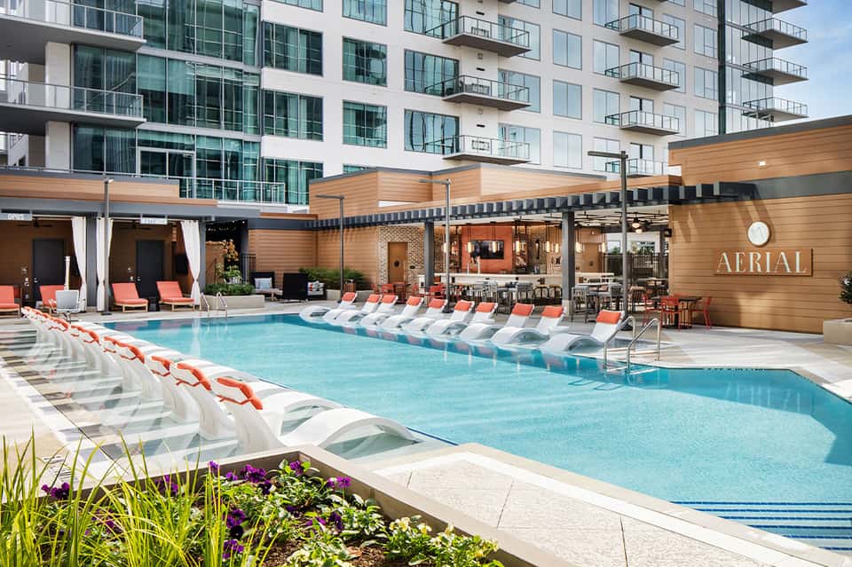 Modern hotel pool deck with loungers, striped umbrellas, wooden pergolas, and contemporary building with balconies