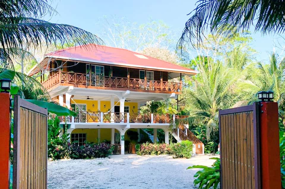 Tropical three-story residential house with colorful yellow and coral exterior, wooden railings, and palm trees