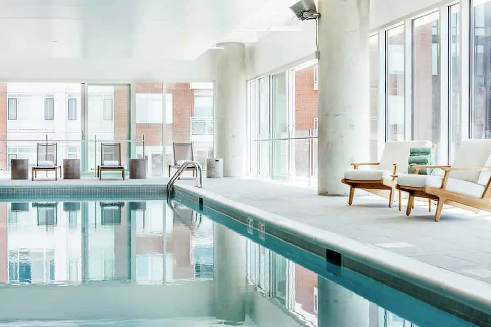Indoor swimming pool with teal edge, wooden lounge chairs, and floor-to-ceiling windows