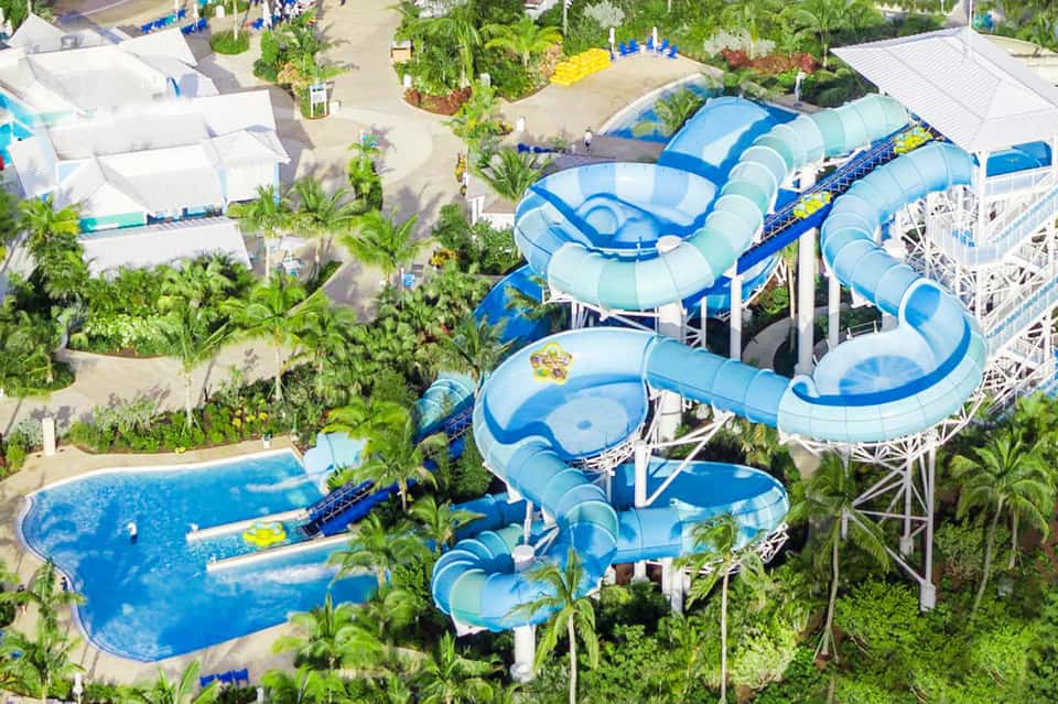 Aerial view of water park with blue twisting slides, pools, palm trees, and white tent structures