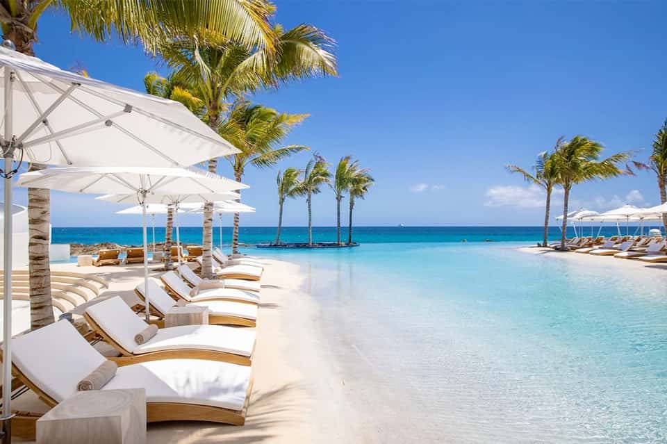 Beachfront infinity pool with white loungers and umbrellas facing turquoise ocean and palm trees