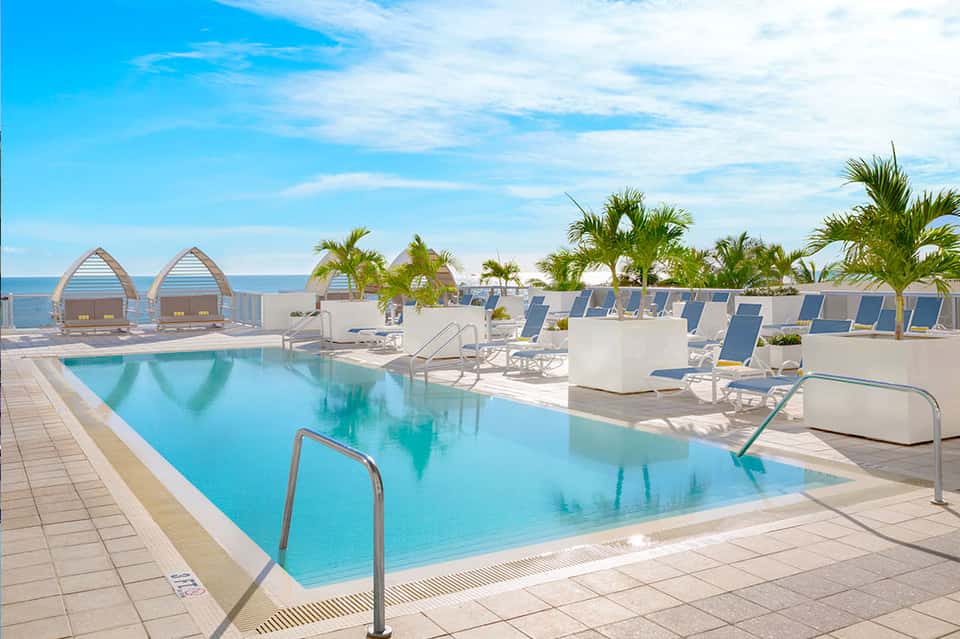 Beachfront resort pool with blue lounge chairs, palm trees, and ocean view cabanas
