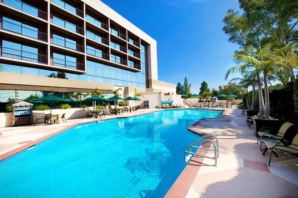 Modern hotel pool with bright blue water, loungers, umbrellas, and multi-story building in background