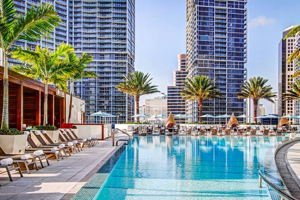 Resort pool deck with loungers, palm trees, and high-rise hotels overlooking waterfront promenade