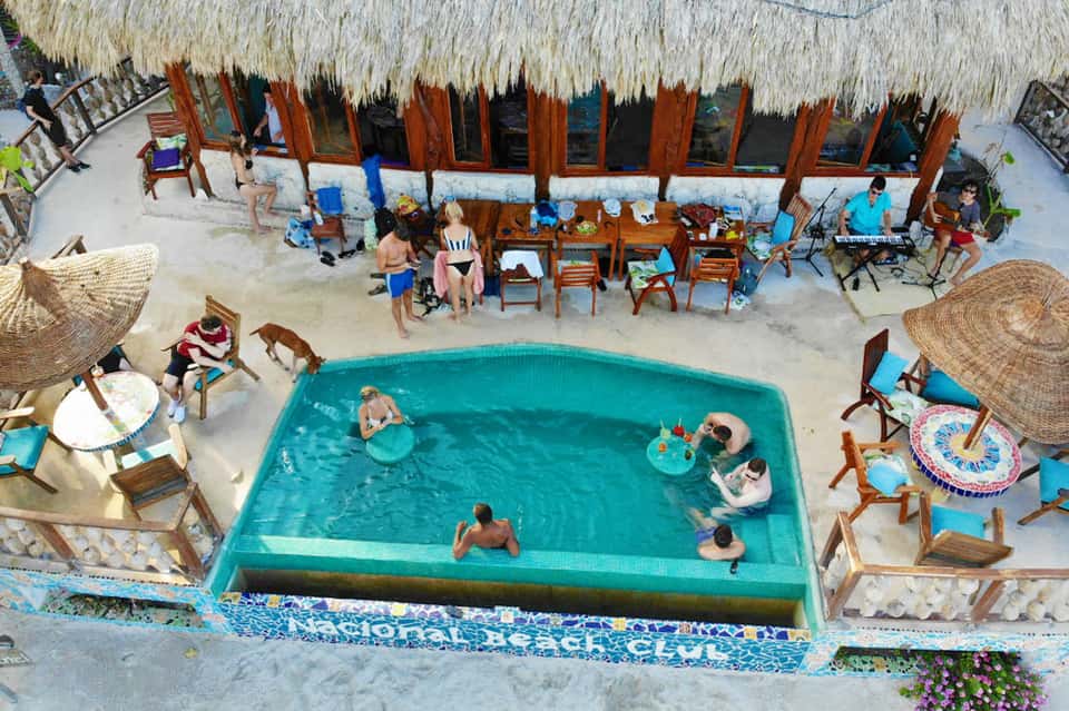 Overhead view of beachside resort pool with thatched-roof cabanas and guests relaxing