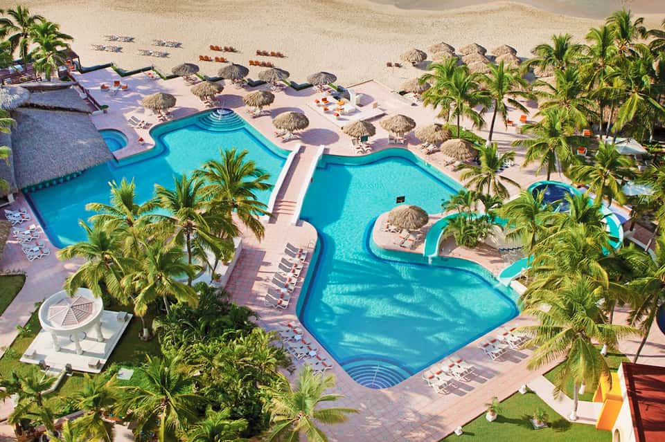Aerial view of resort with curved swimming pools, palm trees, thatched umbrellas, and sandy beach