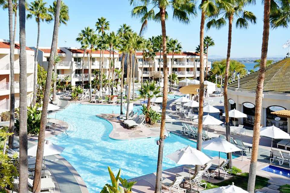 Resort courtyard with curved blue swimming pool, palm trees, and multi-story white buildings under clear sky