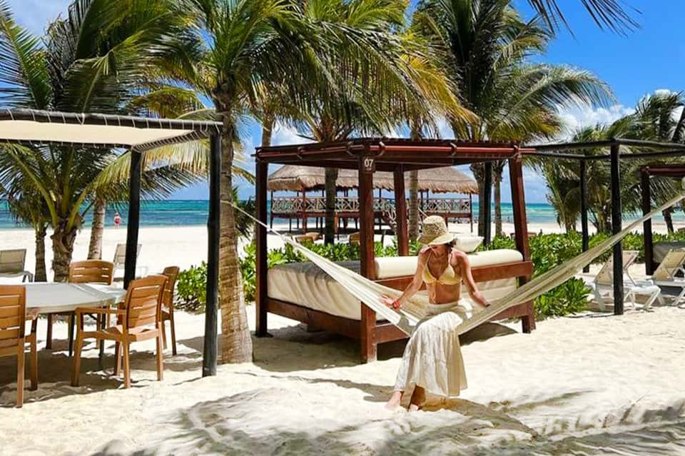 Woman relaxing in hammock on tropical beach with palm trees, ocean view, and thatched-roof structures