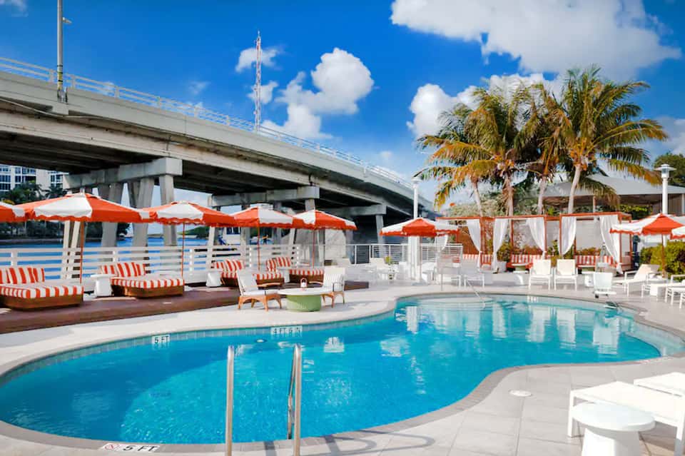 Resort pool with white loungers, orange umbrellas, palm trees under highway bridge