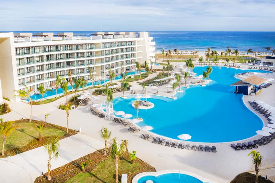 Beachfront resort with expansive curved pool, lounge chairs, white building, ocean and palm trees in background