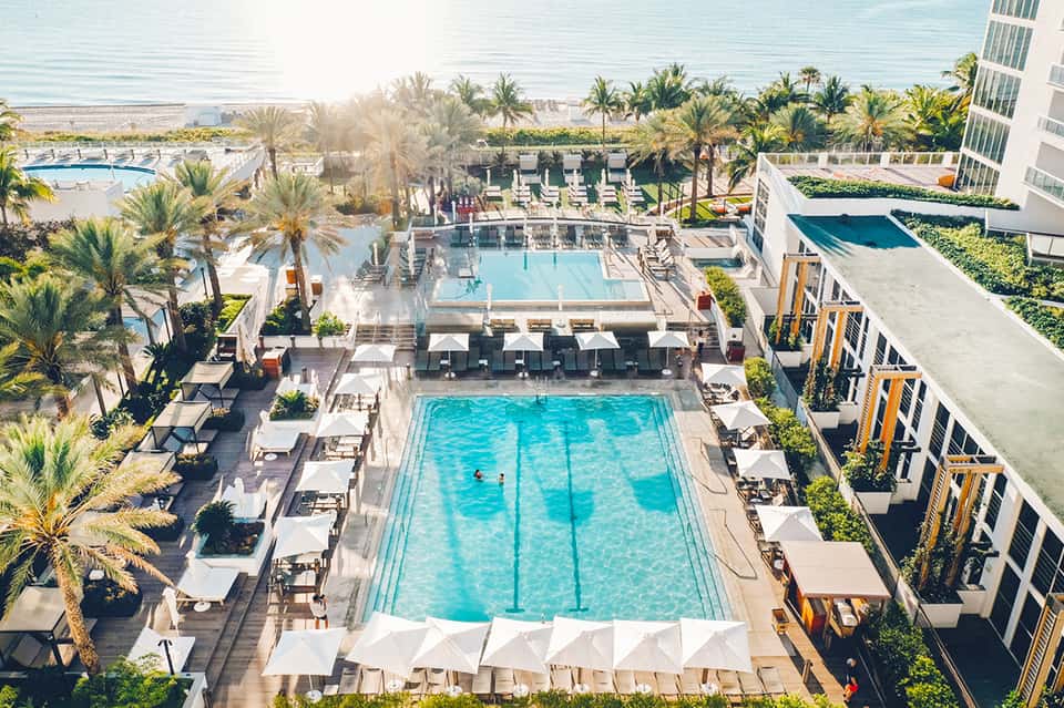 Aerial view of beachfront resort pool complex surrounded by palm trees, loungers, and modern hotel building
