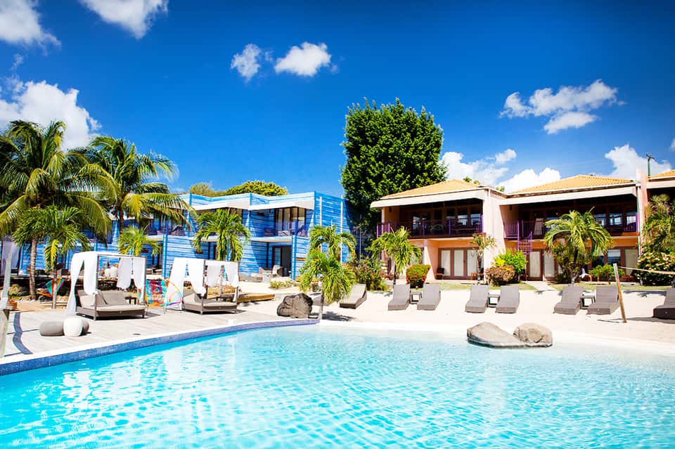 Resort pool with colorful blue and white buildings, palm trees, and lounge chairs under sunny sky
