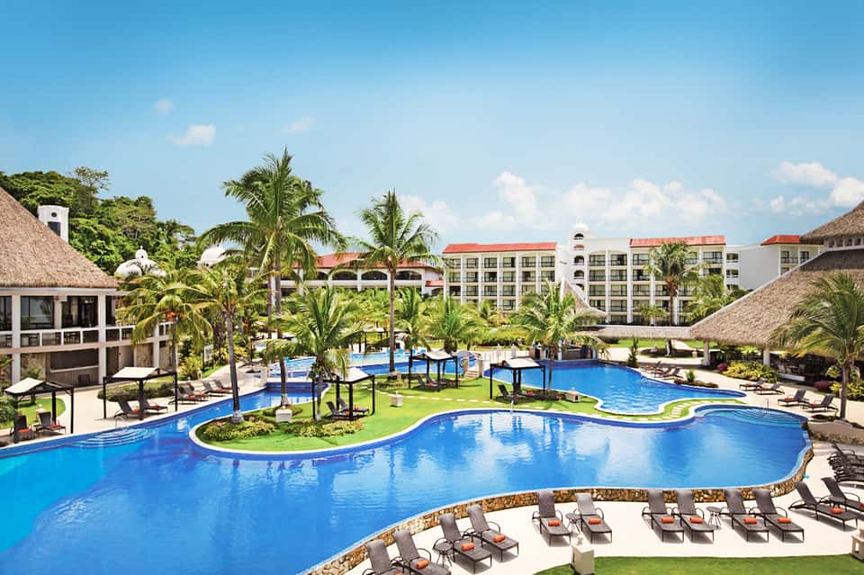 Resort pool area surrounded by white and teal buildings with thatched umbrellas, palm trees, and lounge chairs