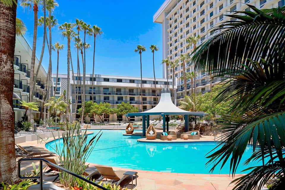 Resort pool with turquoise water, white tents, palm trees, and multi-story hotel buildings