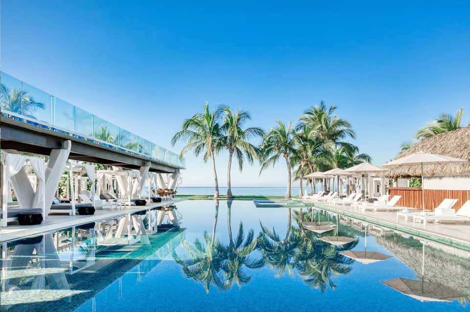 Infinity pool reflecting palm trees and ocean with white cabanas and covered lounging areas