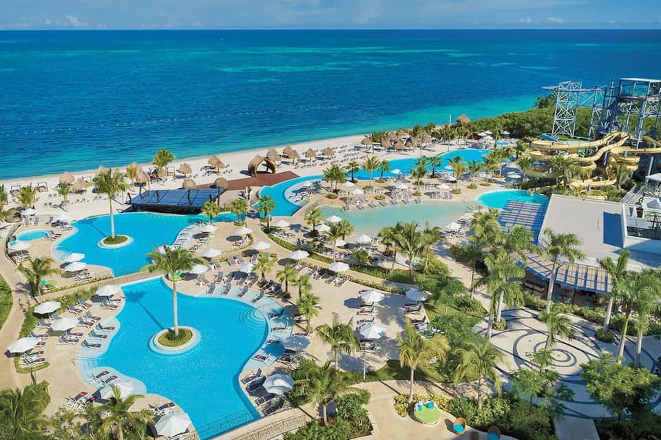 Aerial resort view with blue pools, beachfront, palm trees, and turquoise ocean waters